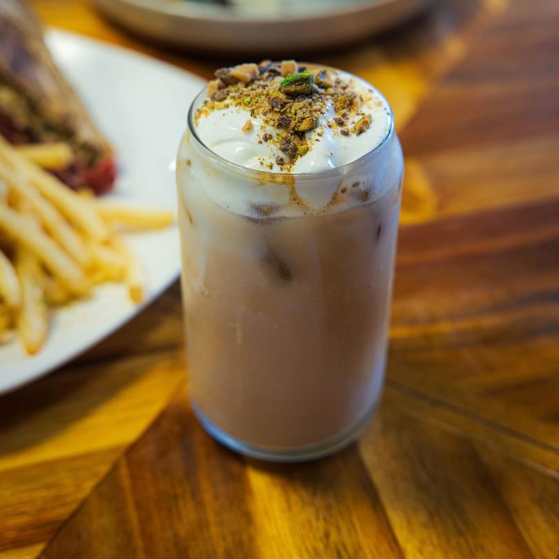 Iced coffee with whipped cream and chocolate crumbles, next to a plate of fries on a wooden table.