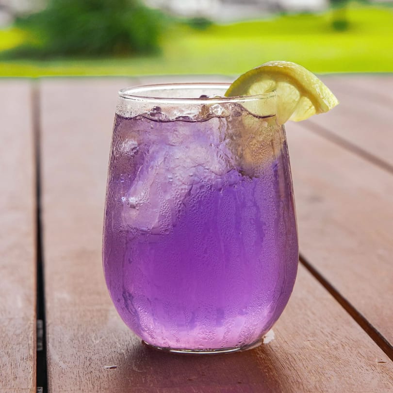 A vibrant purple iced drink with a lemon slice sits on a wooden table outdoors.