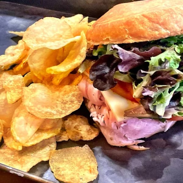 A Meating Ground Cafe sandwich with deli meat, cheese, and lettuce, served with potato chips.